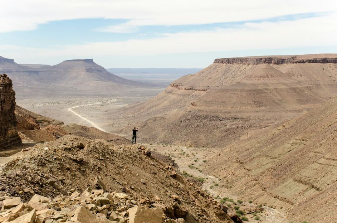person standing at the edge of a rock mountain facing the mountains during day person standing at the edge of a rock mountain facing the mountains during day